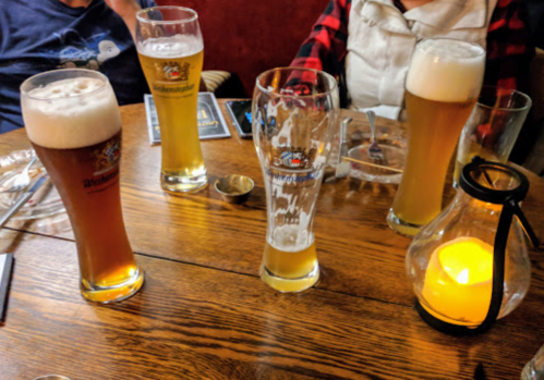 Four glasses of beer on a wooden table, with a lit candle and people seated in the background.