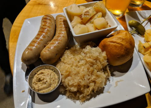 A plate with two sausages, sauerkraut, mustard, a roll, and diced potatoes, served with drinks in the background.