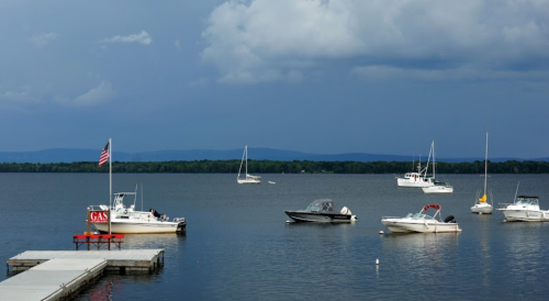 A calm lake scene with various boats anchored, a dock with a gas sign, and dark clouds in the background.