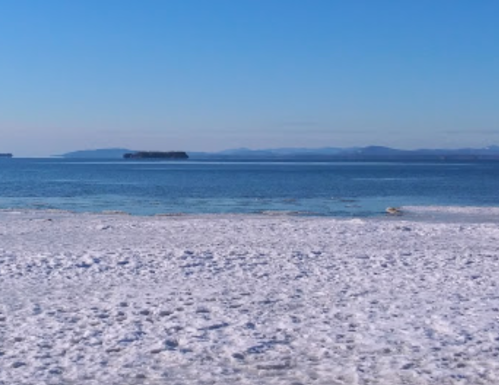 A serene winter beach scene with snow-covered sand and calm blue water under a clear sky.