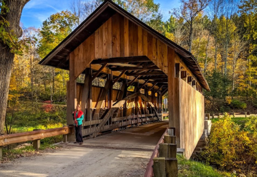 A person stands near a wooden covered bridge surrounded by autumn foliage and trees.