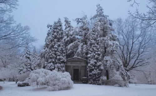 A snow-covered mausoleum surrounded by tall evergreen trees in a serene winter landscape.