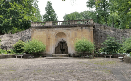 A stone wall with a statue in a serene garden setting, surrounded by greenery and benches.