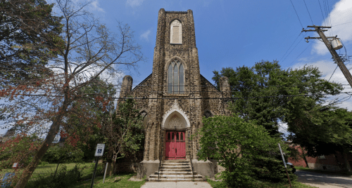 Gothic-style stone church with a tall tower, red doors, and surrounding greenery under a blue sky.