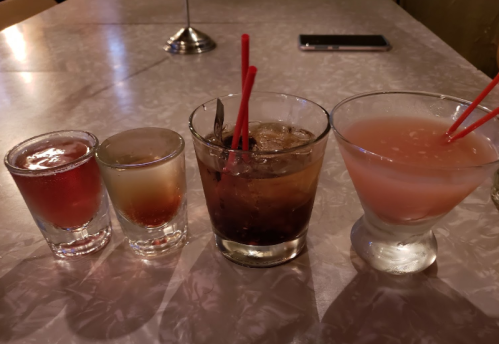 Four colorful cocktails on a table, including shots and mixed drinks, with straws and a phone in the background.