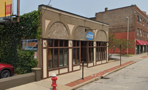 A street view of a restaurant with arched windows and a sign reading "Side Quest," surrounded by greenery and buildings.