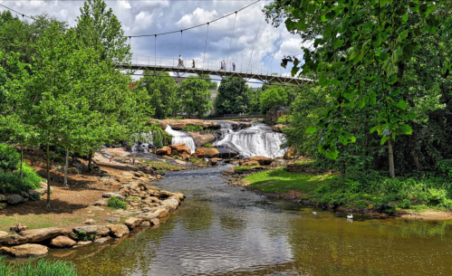 A scenic view of a river with a waterfall, surrounded by greenery and a pedestrian bridge in the background.