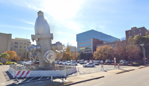 A large, metallic fire hydrant sculpture stands in a parking lot, surrounded by buildings and trees.
