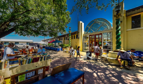 A vibrant outdoor market scene with people browsing books, colorful stalls, and a sunny blue sky.
