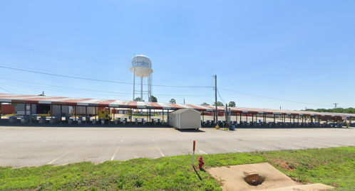 A wide view of a parking lot with a covered structure and a water tower in the background under a clear blue sky.