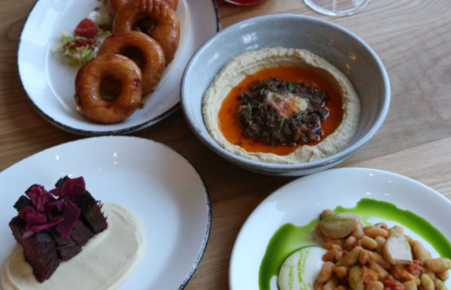 A spread of dishes including hummus with toppings, fried doughnuts, a meat dish, and a bean salad on white plates.