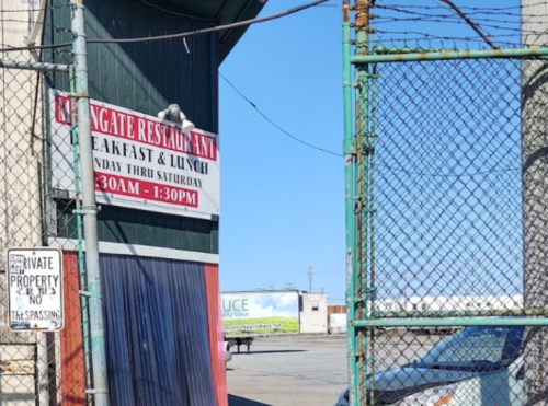 Sign for "Langate Restaurant" with hours, framed by a fence and a view of a parking area. Clear blue sky in the background.