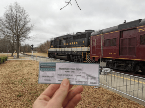 A hand holds a train ticket in front of a passing train at a station, with trees and a fence in the background.