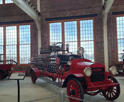 A vintage red fire truck displayed indoors, with large windows in the background and historical signage nearby.