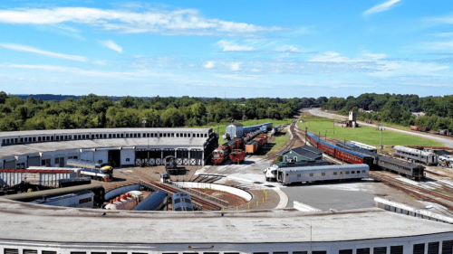 A panoramic view of a train yard with multiple tracks, trains, and green fields under a blue sky.
