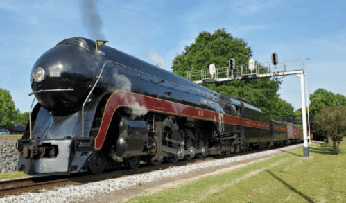 A vintage steam locomotive with a shiny black exterior and red accents, passing under a signal bridge on a sunny day.