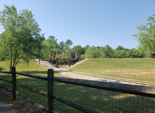 A wooden bridge spans a grassy area with train tracks, surrounded by trees under a clear blue sky.