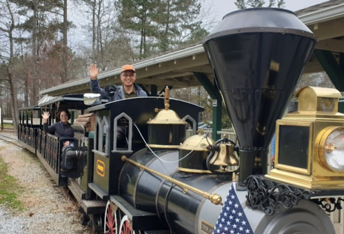 A vintage-style train with passengers waving, set in a park with trees in the background.