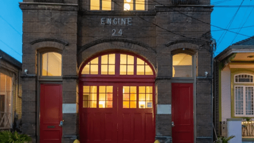 Historic fire station with red doors and large windows, illuminated at dusk.