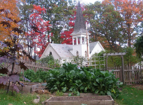 A quaint church with a tall spire surrounded by vibrant autumn foliage and a lush vegetable garden in the foreground.