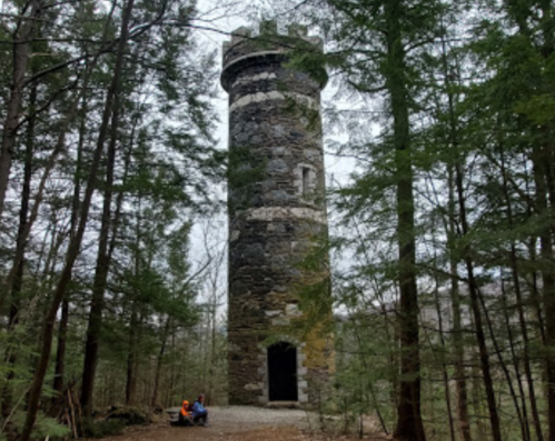 A stone tower surrounded by trees, with two people sitting on the ground nearby in a forested area.