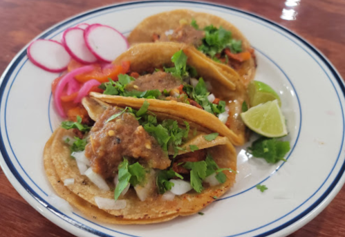 Three tacos topped with meat, onions, and cilantro, served with lime wedges and pickled radishes on a white plate.