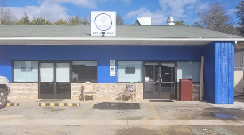 A blue building with a sign, featuring two chairs outside and a parking area in front.