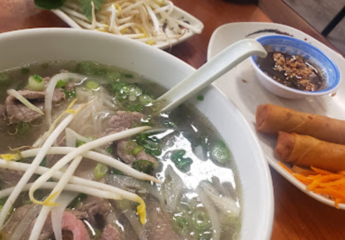 A bowl of pho with beef, bean sprouts, and herbs, alongside spring rolls and a small dish of dipping sauce.