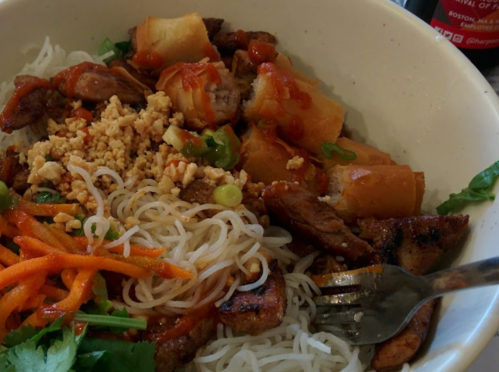 A bowl of noodles topped with grilled meat, crispy vegetables, peanuts, and a drizzle of sauce, with a fork beside it.