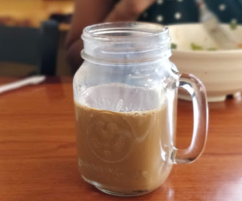 A glass mason jar filled with brown coffee sits on a wooden table, with a blurred background of a bowl and a person.