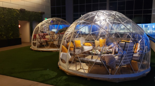 Two transparent igloo-shaped structures with cozy seating and yellow cushions, set on green grass indoors.