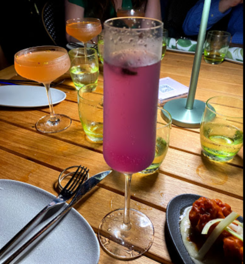A pink cocktail in a tall glass sits on a wooden table surrounded by various drinks and plates.