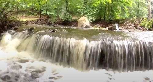 A serene waterfall cascades over rocks, surrounded by lush green trees in a peaceful natural setting.