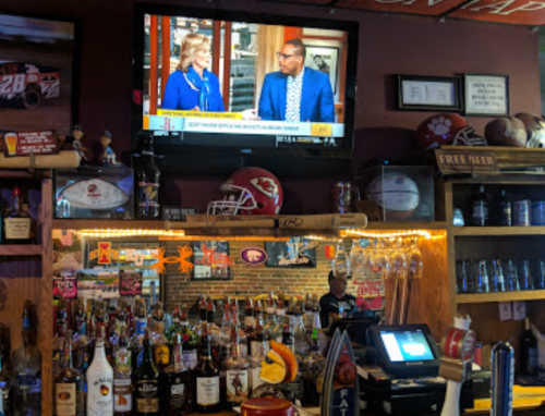 A bar scene with a TV showing a news program, shelves of liquor bottles, and a bartender in the background.