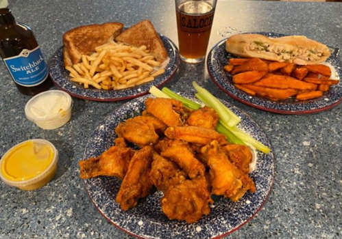A table spread with fried chicken wings, sweet potato fries, toast, a sandwich, and a beer.
