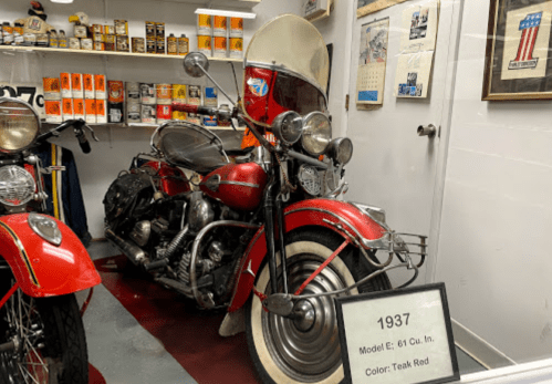 A vintage 1937 motorcycle, Model E, 61 Cu. In., in teak red, displayed in a museum with various memorabilia in the background.