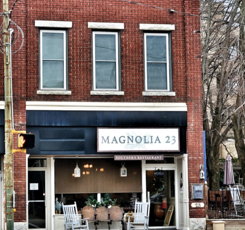 Exterior of Magnolia 23, a Southern restaurant, featuring a brick facade and two rocking chairs on the porch.