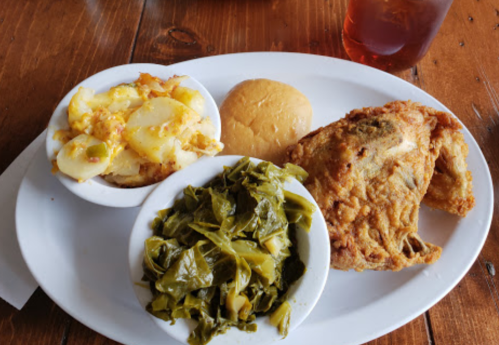 A plate with fried fish, collard greens, cheesy potatoes, and a bun, served on a wooden table with a drink.