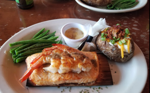 A plate with grilled salmon topped with shrimp, green beans, and a loaded baked potato with cheese and bacon.