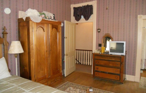 A vintage bedroom featuring a wooden wardrobe, dresser, and a small TV, with patterned wallpaper and a doorway.