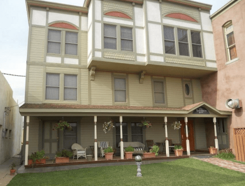 A two-story building with a porch, large windows, and potted plants in front, set in a sunny outdoor area.