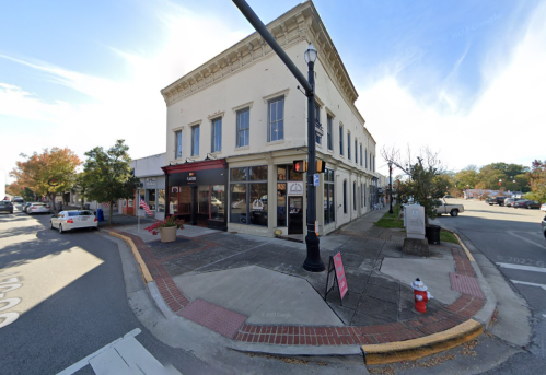 A historic building on a street corner with a traffic light, trees, and parked cars in a small town setting.