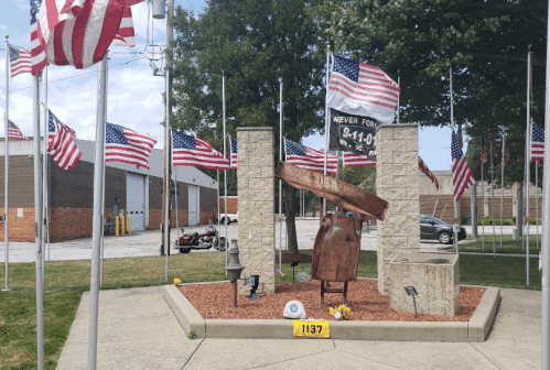 A memorial with flags, a bell, and stone pillars, honoring those who served, surrounded by a landscaped area.