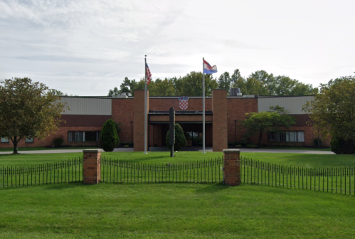 A brick building with flags in front, surrounded by green grass and trees, under a cloudy sky.