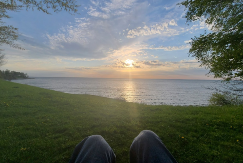 A serene view of a sunset over a calm lake, with grassy foreground and a cloudy sky.