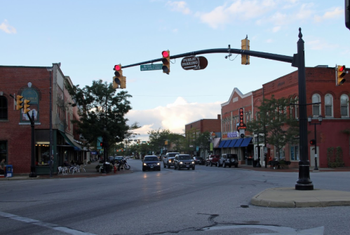 A street view of a small town with shops, traffic lights, and cars under a partly cloudy sky.