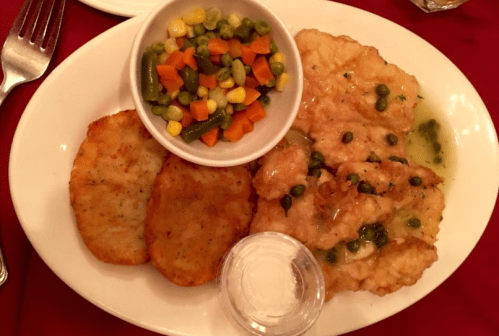 A plate with fried fish topped with green sauce, two potato cakes, and a side of mixed vegetables.