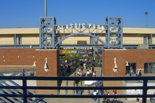 Entrance to Classic Park with people walking in on a sunny day, surrounded by blue skies and stadium structures.