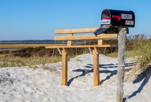 A wooden bench beside a black mailbox labeled "Kindred Spirit" on a sandy beach with grass and water in the background.