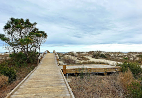 A wooden boardwalk leads through sandy terrain to the beach, with sparse vegetation and a cloudy sky above.